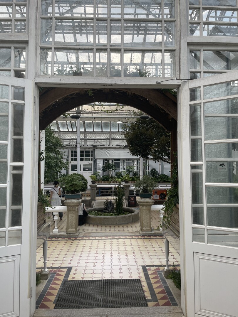 The entrance to a greenhouse built in 1909. in the Center of the greenhouse is a small pond with some small koi. Around the pond are some larger trees and along the walls of the greenhouse a collection of various bonsai.