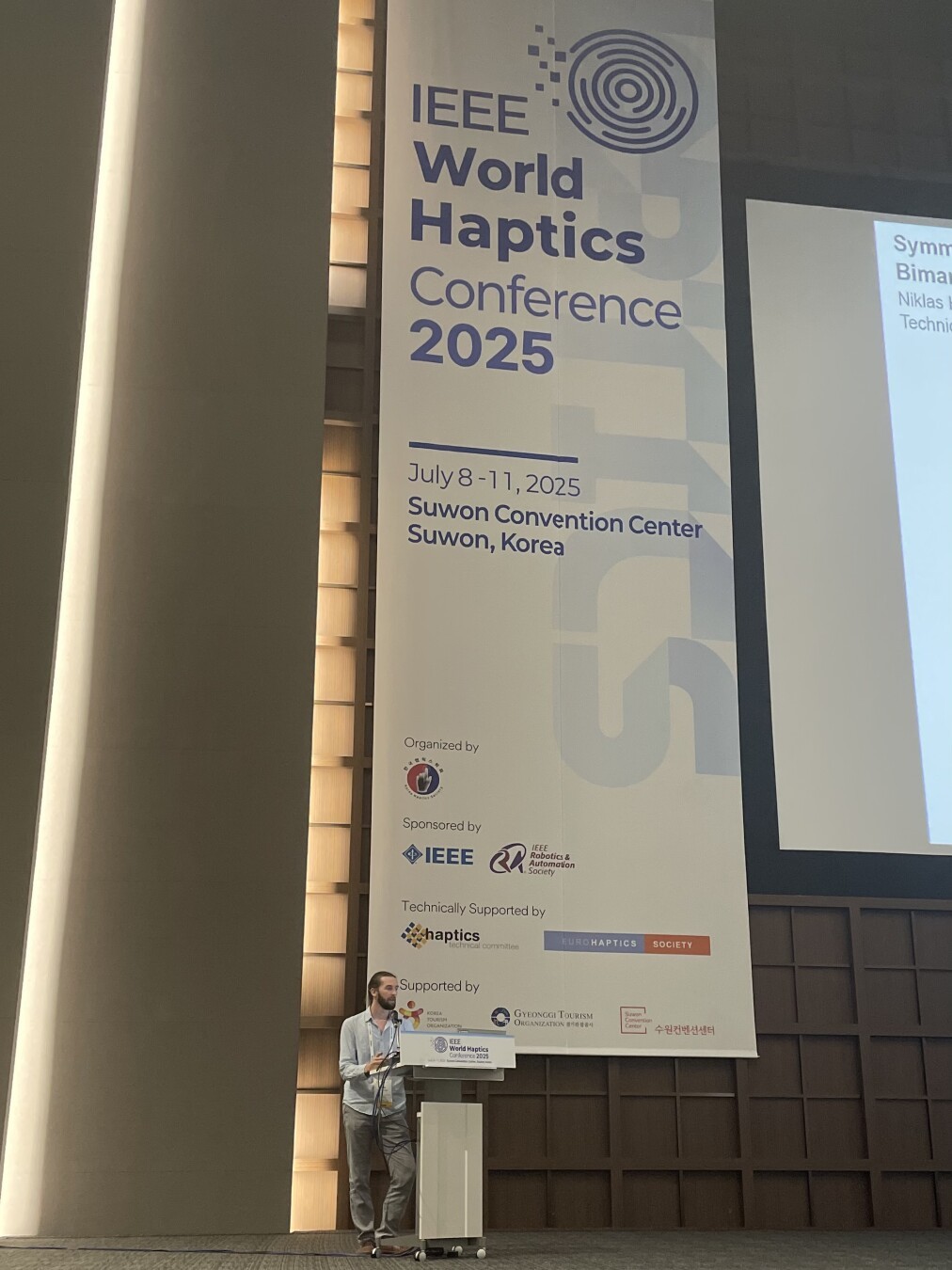 Me on stage at World Haptics 2025 presenting a teaser for my research in front of a big world haptics banner.