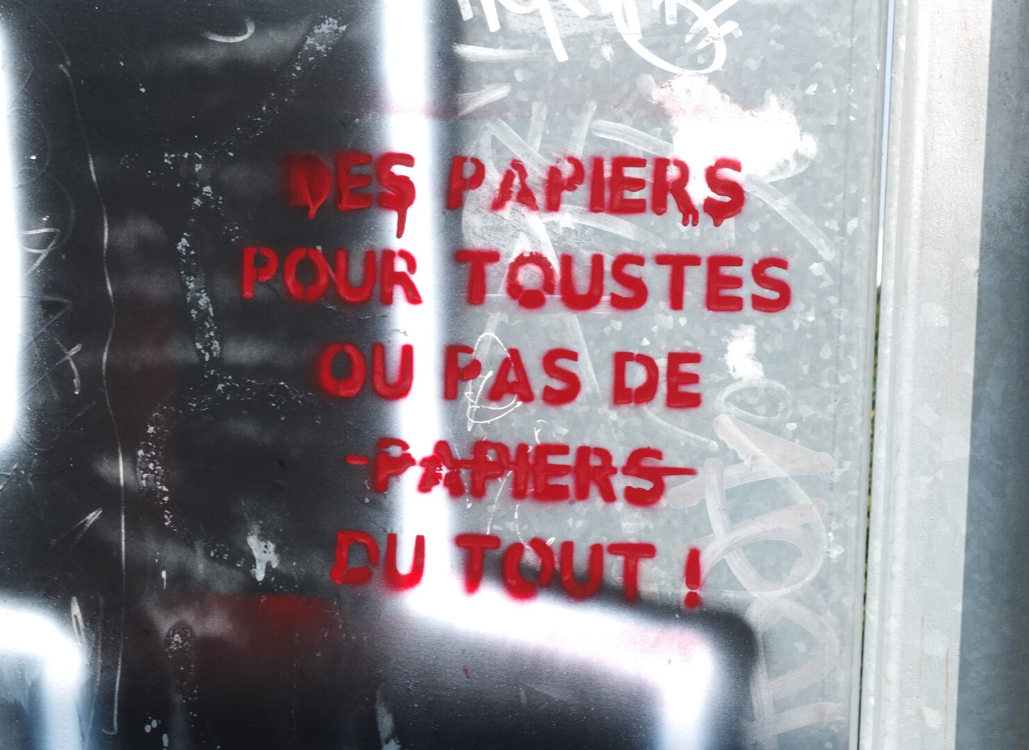 Red text painted on metal : "Des papiers pour toustes ou pas de papiers du tout" (Papers for everyone or no papers at all)