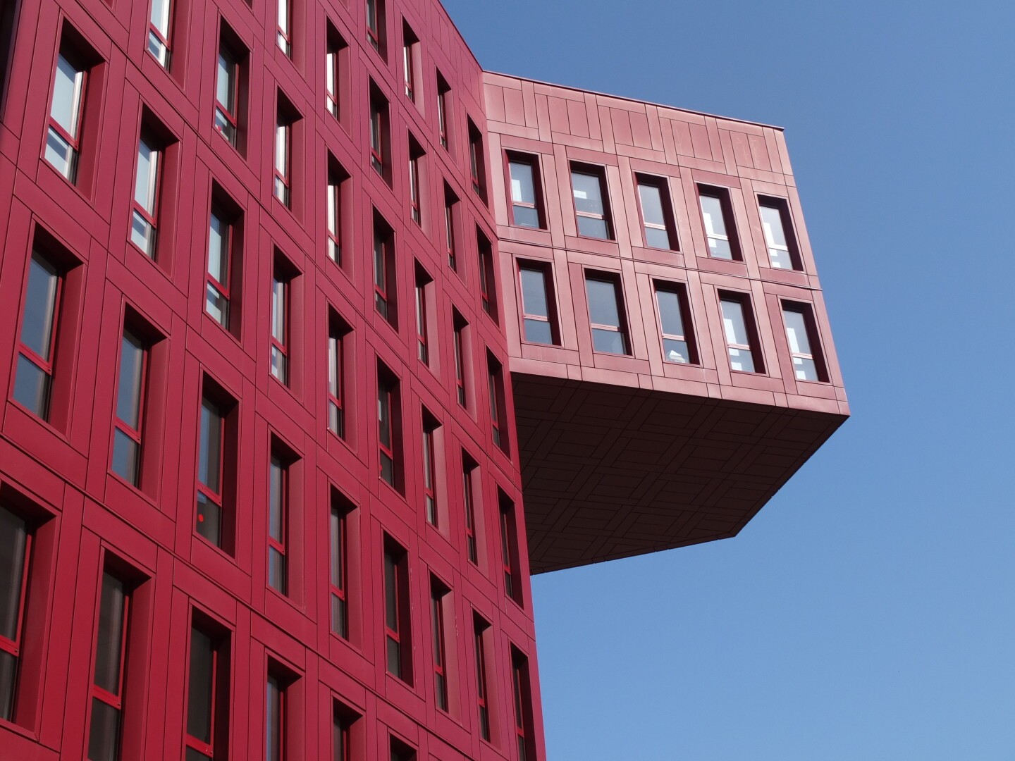 Modern red building with rectangular windows, featuring a large cantilevered section extending outward against a clear blue sky