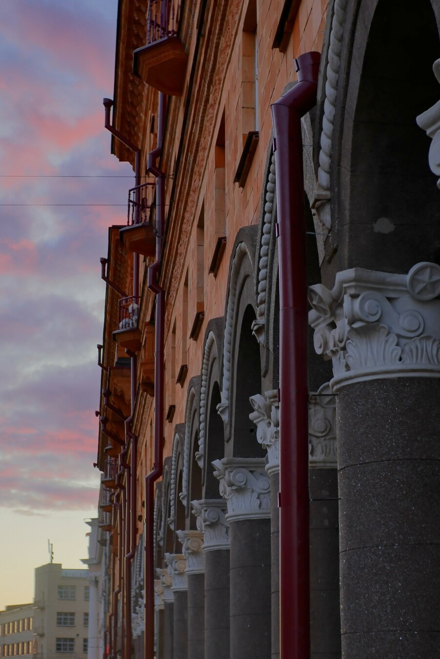 A close-up, low-angle view of a Stalinist-era building on Praspiekt Niezaležnasci in Minsk, Belarus, at the beginning of a sunset.
A row of arches is supported by columns with decorative white capitals, and red downspouts running vertically along the building.
The building's warm, orange-toned stone or brick contrasts with the grey stone arches and the pastel-colored sky in the background