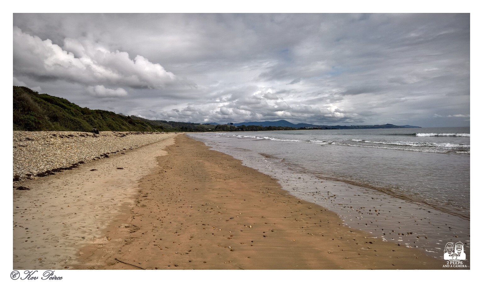 A wide, horizontal photo of a sandy beach stretching into the distance under a dramatic, cloudy grey sky.

To the left, the beach meets a bank covered in rough stones and scrubby green vegetation.

The sand is wet and reflective where it meets the incoming gentle waves on the right. In the distance, a coastline and rolling hills or mountains are visible under the clouds.

The photo is signed 'Kev Peirce'.