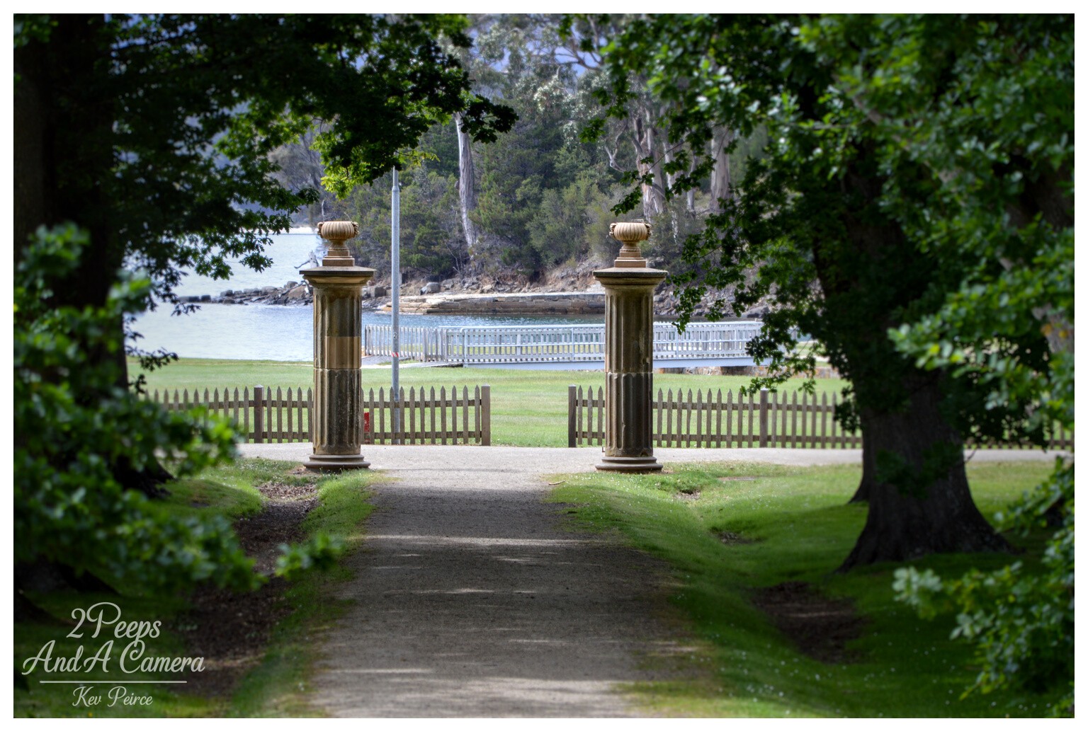 A slightly shaded, gravel path framed by large trees leads through an open gate with two classical stone columns toward a grassy lawn, a picket fence, and the calm blue water of the bay at Port Arthur.

Photo by Kev Peirce.