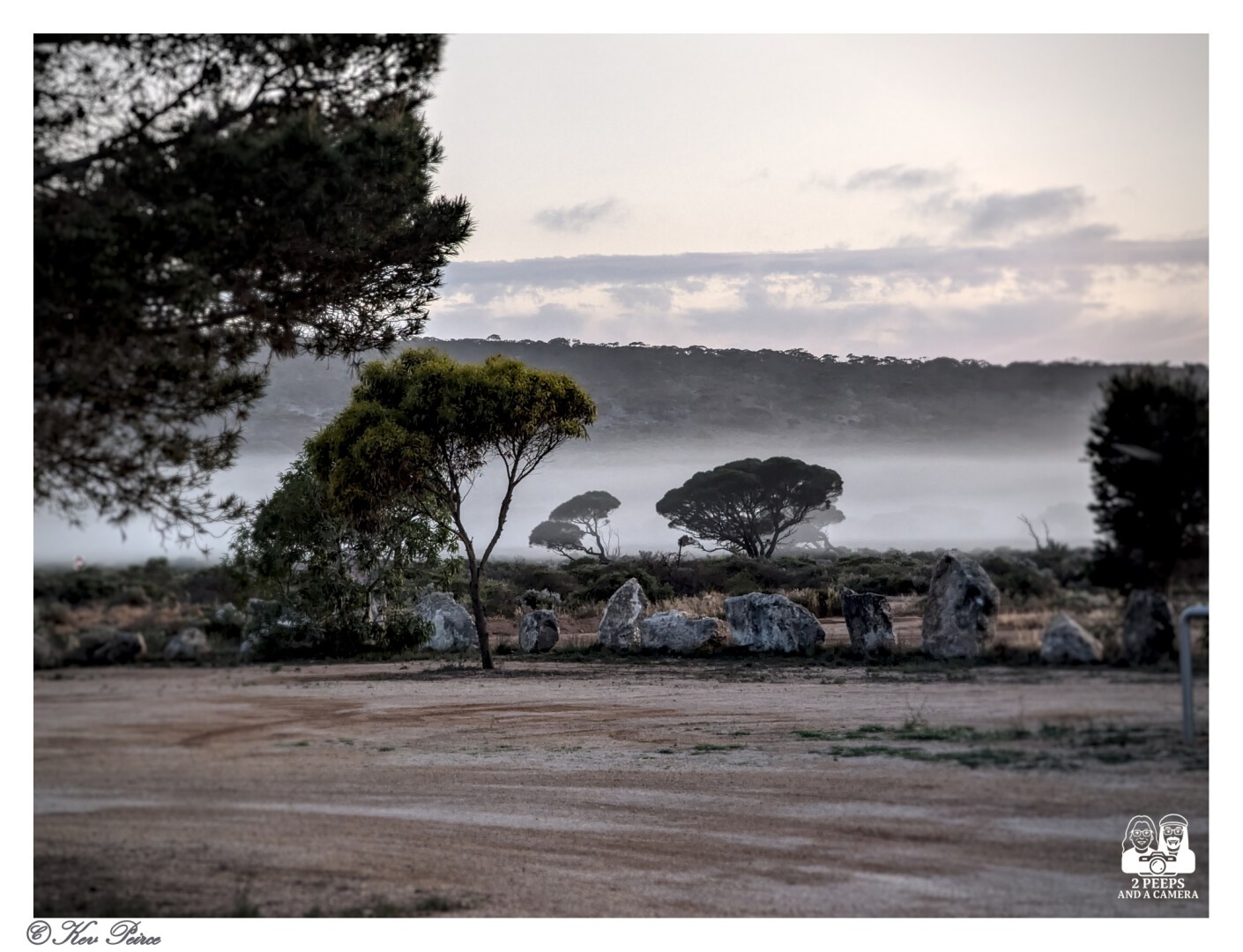 A moody landscape photograph near Mundrabilla. Mist or fog hangs low across the middle ground, obscuring the base of several dark, solitary trees and a line of white rocks.

The foreground consists of dirt and dry ground, and the background shows a dark, wooded ridge under a pale, overcast sky.