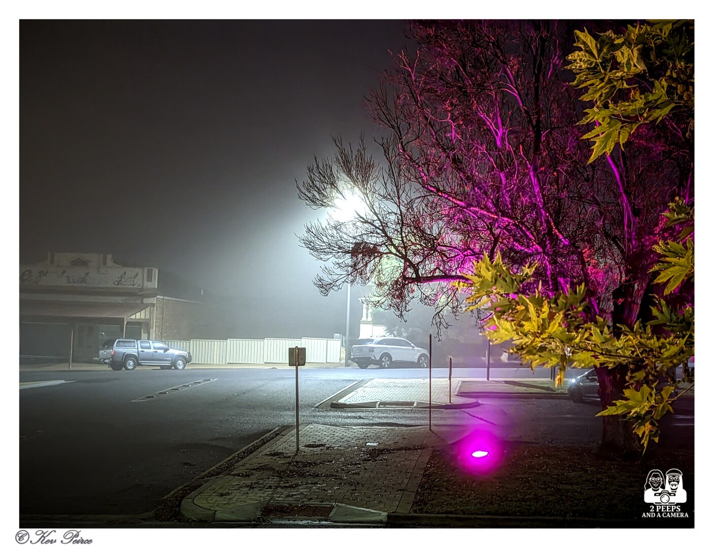 A moody, atmospheric photo of an empty street and parking lot in Loxton, Australia, on a foggy night. A very bright streetlight in the center is surrounded by a foggy halo.

The bare branches of a large tree on the right are illuminated by a vibrant, unnatural pink and purple light, contrasting with the yellowish leaves on the right edge.

A few parked cars are visible on the left and center-right. Signed by Kev Peirce.