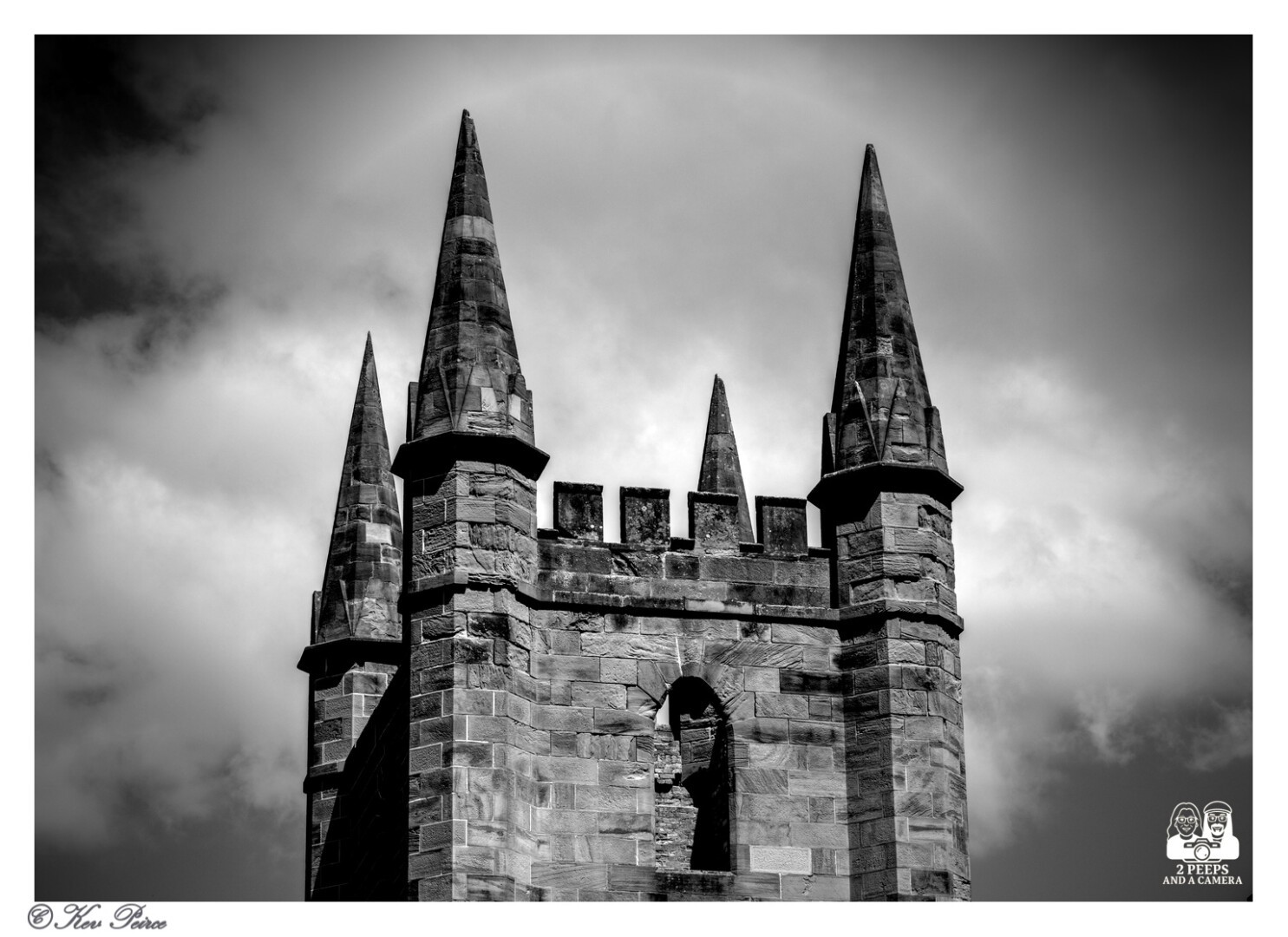 A dramatic black and white photograph of the top of the ruined Anglican Church at Port Arthur, Tasmania.

The stone tower, with its distinctive spire-like pinnacles and crenellated top, is silhouetted against a cloudy, bright sky, creating a high-contrast and moody effect.

A halo effect is visible around the main structure against the clouds. The photograph is signed by Kev Peirce.
