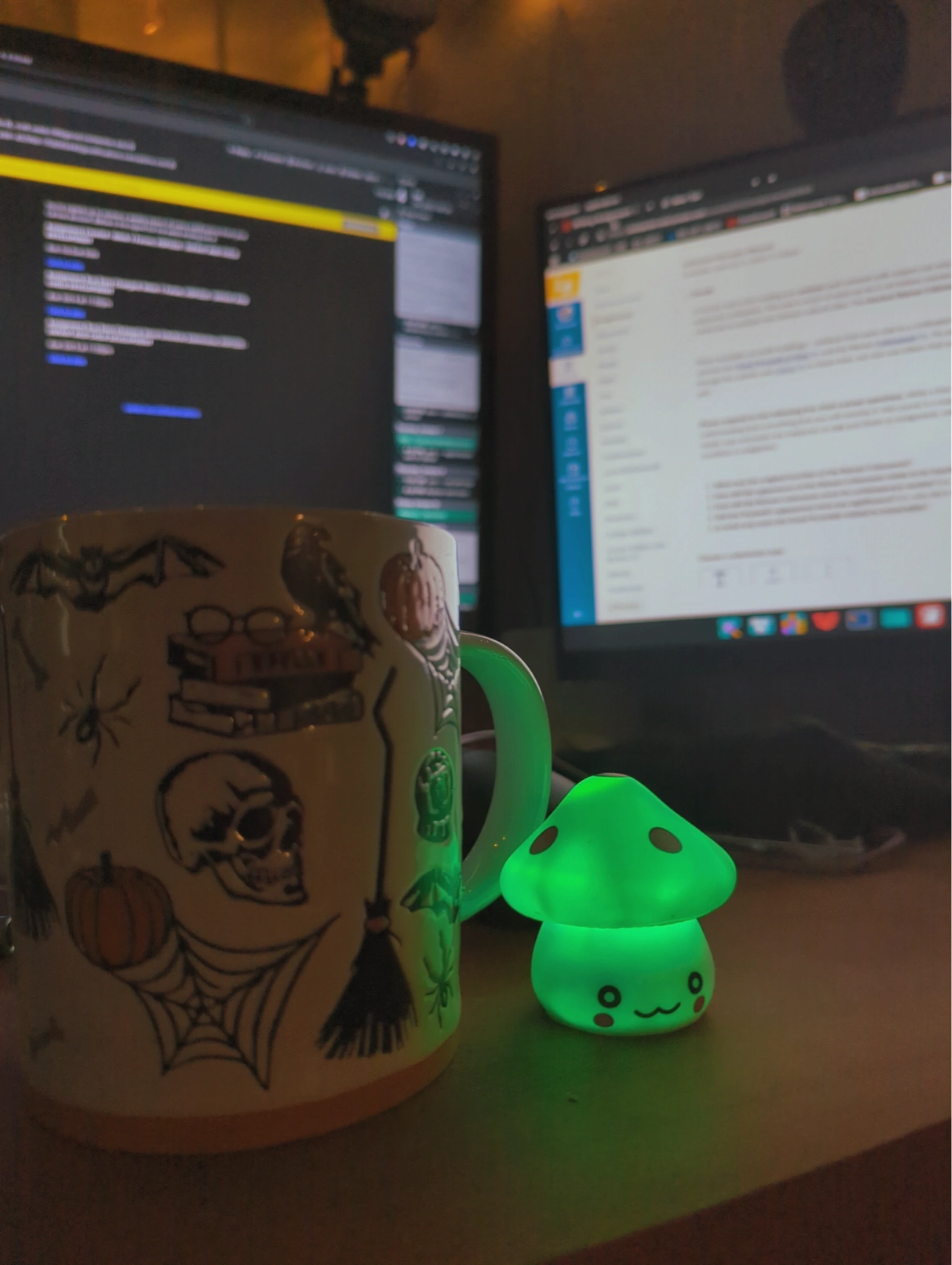 A coffee cup with spooky Halloween themed items such as bats, a skull, and spider webs is sitting next to a battery powered mushroom that lights up green. The background is slightly blurred where computer monitors are on.