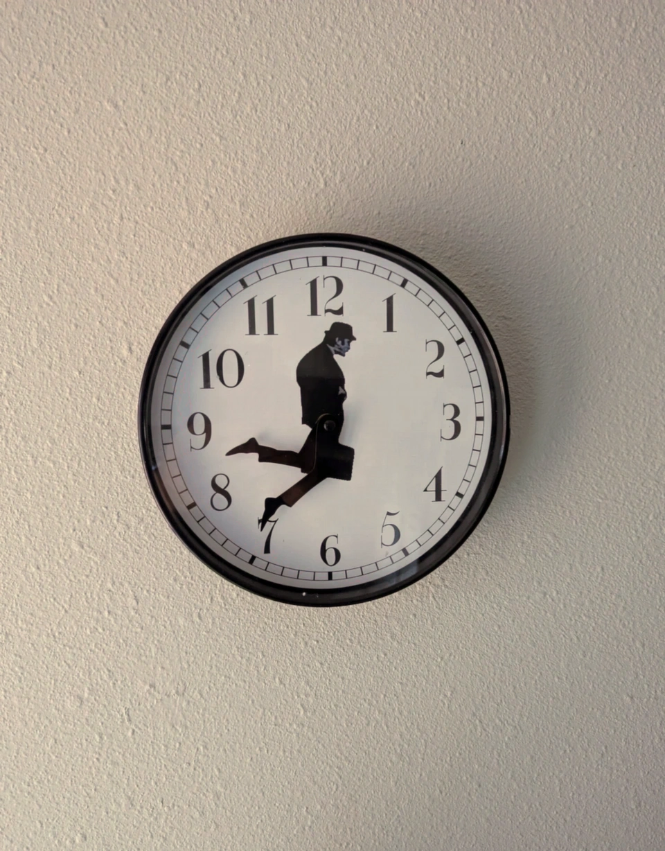 A circular, black-framed wall clock that has a white background. John Cleese from the comedy show Monty Python is the center piece of the clock, where it's a side profile of his body in which his legs can point at the hours and minutes on the clock face. He is wearing the outfit he wore in the Ministry of Silly Walks.