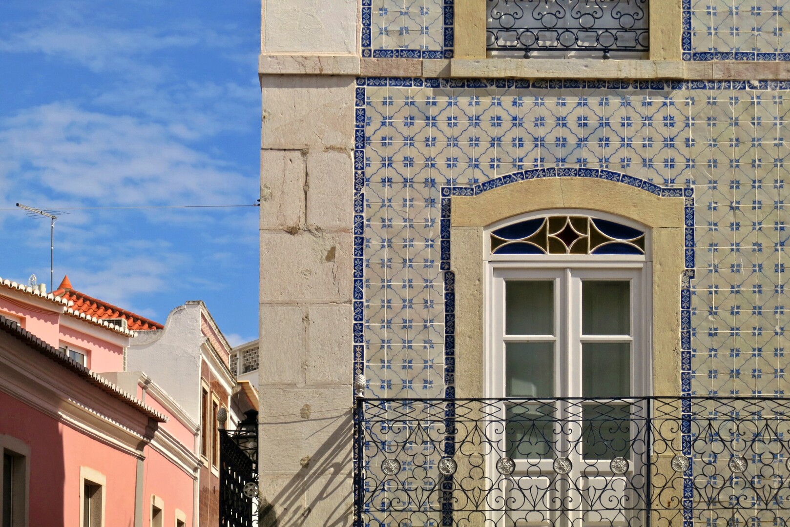 Die Frontalansicht eines Altbaus nimmt zwei Drittel des Bildes ein. Man sieht ein Fenster mit weißem Rahmen, davor das filigran geschmiedet Geländer eines Balkons. Die Fassade ist mit weiß-blau gemusterten Keramikfliesen bedeckt. Im linken Bilddrittel sieht man eine Straßenflucht mit rosafarbenen Fassaden. Darüber der hellblaue Sommerhimmel.