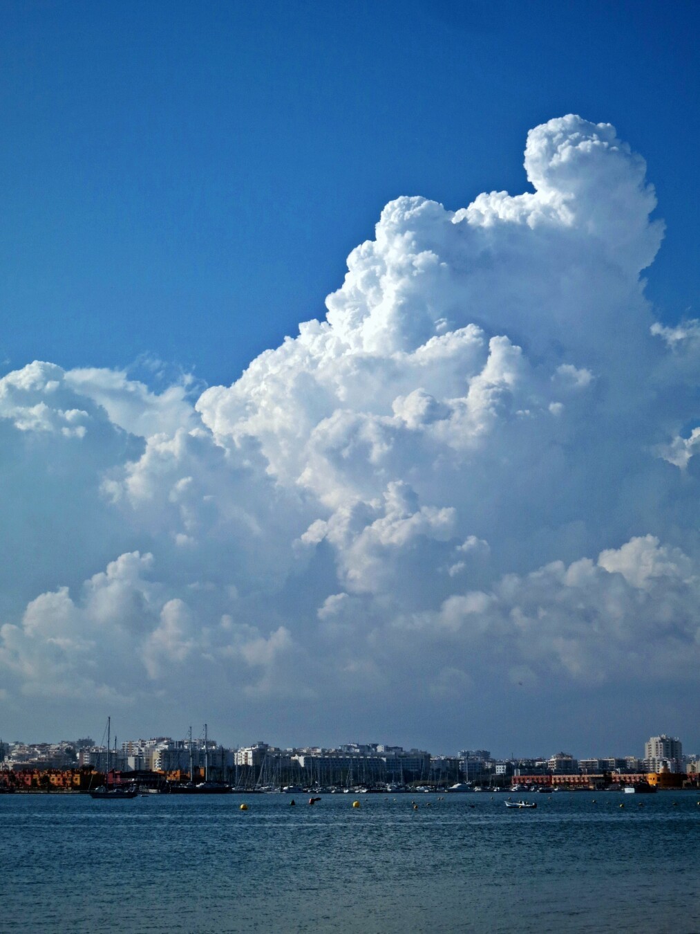 Eine riesige Cumuluswolke nimmt etwa 4/5 der Bildfläche ein. Unter ihr ist die Hafenansicht einer Stadt zu sehen, die im Vergleich winzig wirkt. Davor liegt das Meer.