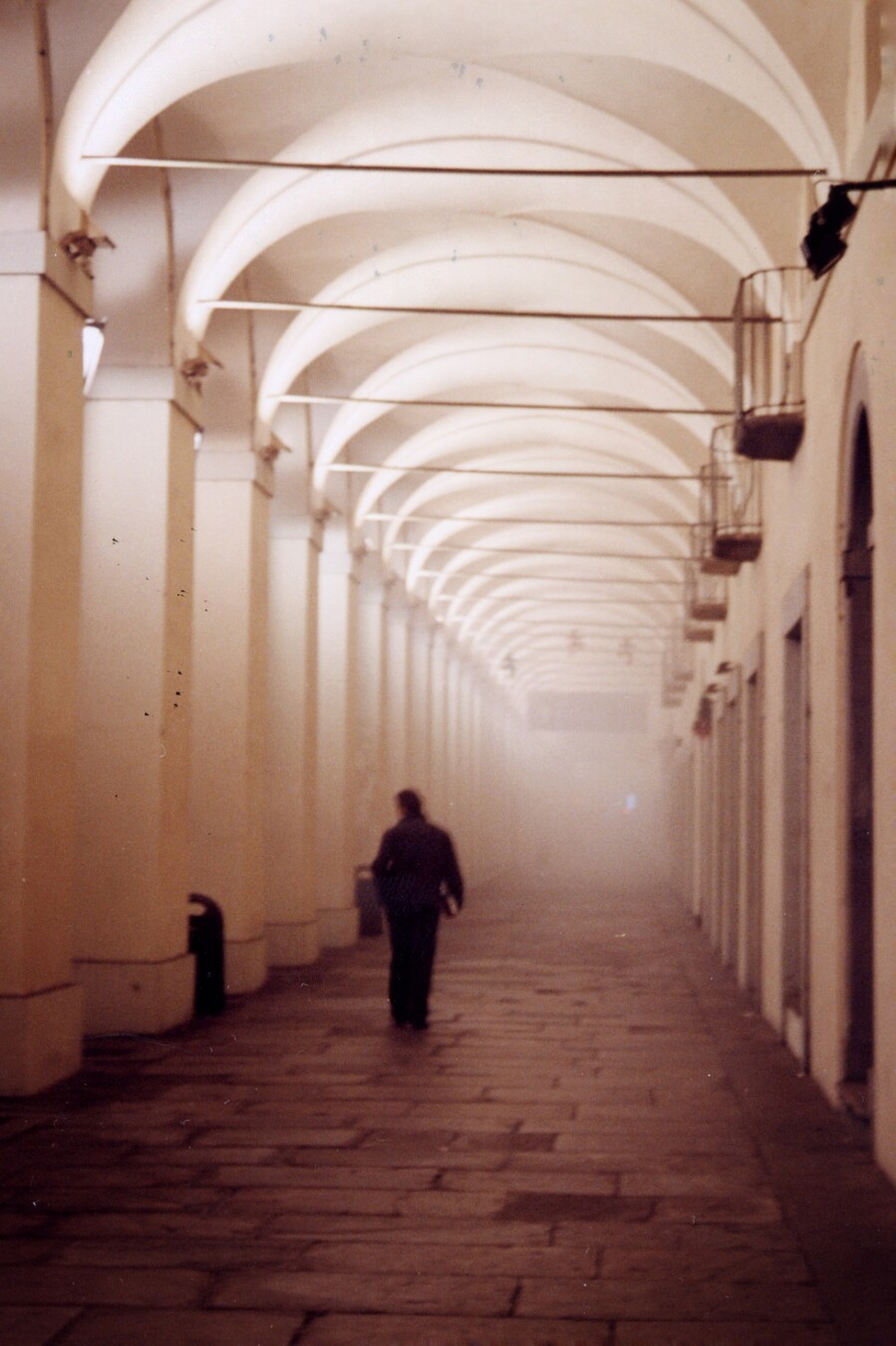 Ein scheinbar endloser Arkadengang im Nebel. Darin ist ein schreitender Mann als Silhouette zu erkennen, von hinten im Gegenlicht. Er geht auf die Nebelwand zu und wird darin verschwinden.