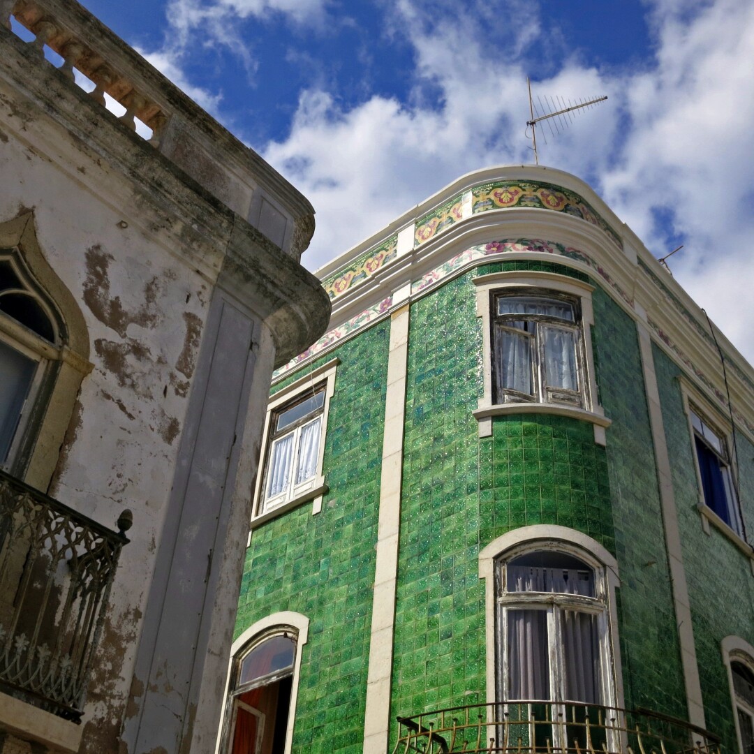 Zwei alte Gebäude sind in Untersicht zu sehen. Die Linien ihrer Fassaden fluchten steil zum bewölkten Sommerhimmel hin. Vom Gebäude im Vordergrund ragt nur eine Ecke ins Bild. Der ehemals weiße Putz ist in die Jahre gekommen und von braunen Flecken bedeckt. Die smaragdgrüne Keramikfassade eines hinteren Gebäudes dagegen schimmert im Sonnenlicht. Die Attika ist mit floralen Mustern verziert.