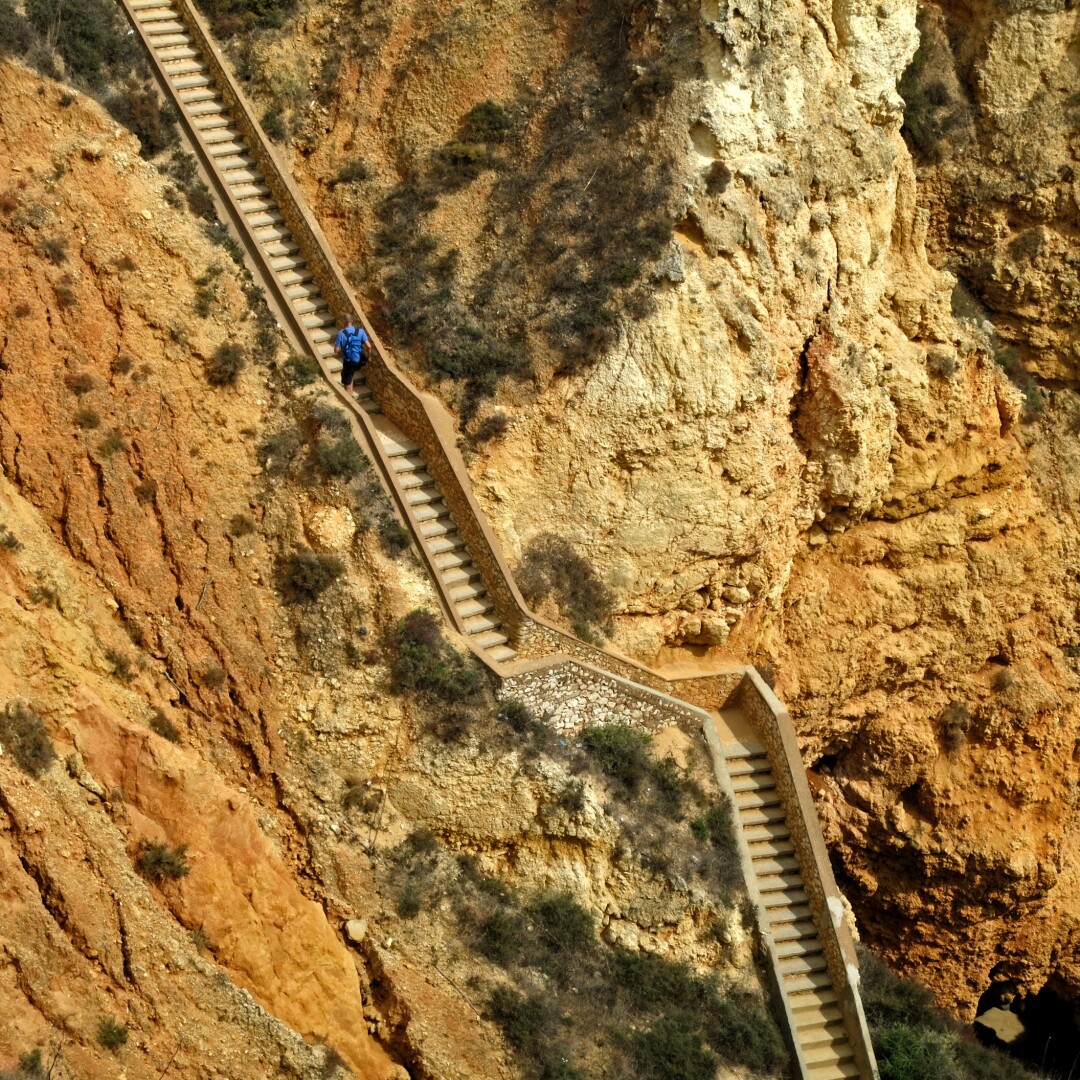 Eine schroffe Klippe in Ocker und Sandfarben. Diagonal durchs Bild verläuft eine steile Treppe, die in den Fels gebaut wurde. Darauf sieht man einen Menschen, der die Treppe hinaufsteigt und im Verhältnis winzig erscheint.