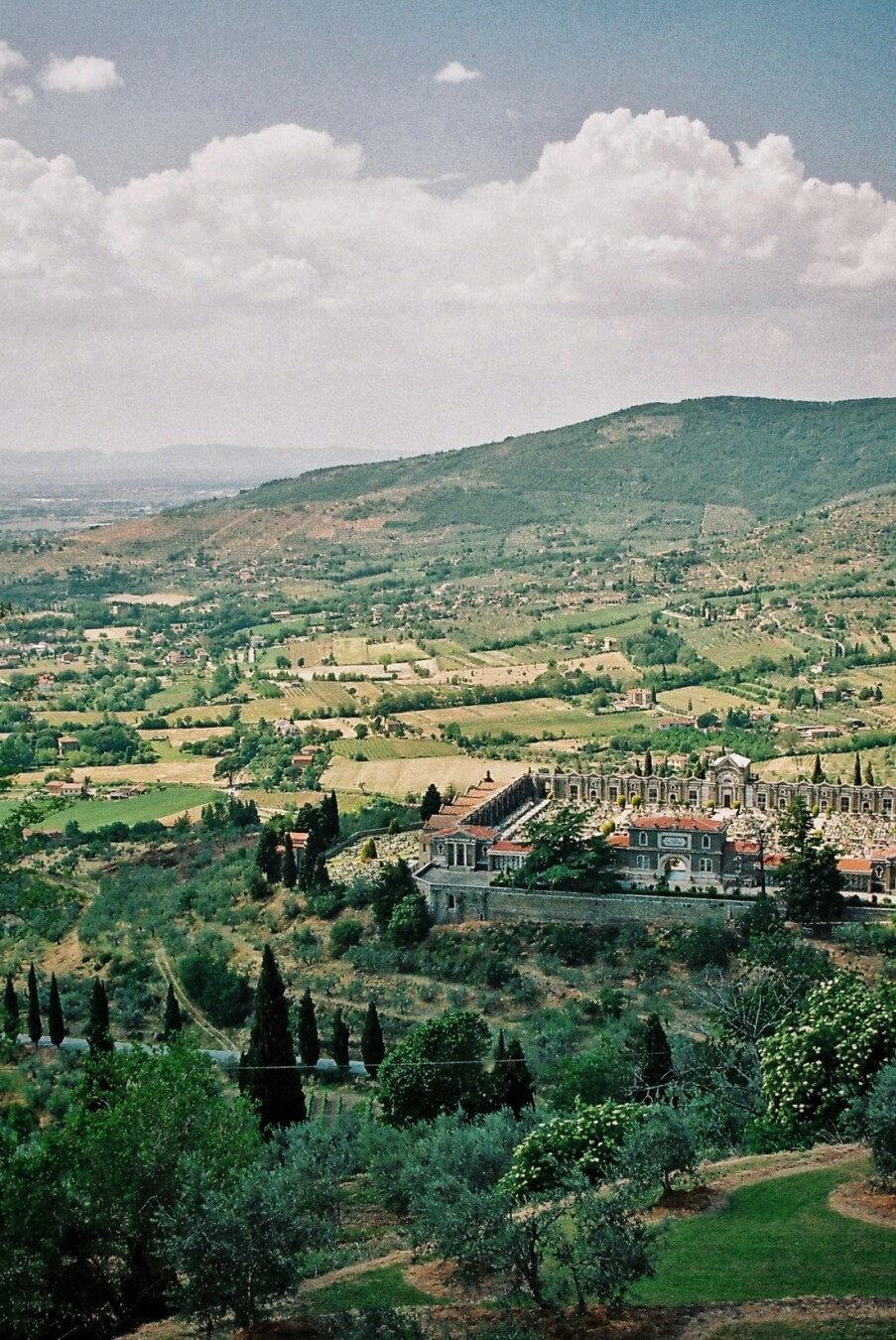 Blick über eine Landschaft. Das obere Bilddrittel ist vom Sommerhimmel eingenommen, über den Cumuluswolken ziehen. Darunter erstrecken sich sanfte grüne Hügel  der Toskana. Rechts in der Mitte des Bildes ist ein alter Friedhof zu sehen. Er ist von einer Mauer umgeben, die einen annähernd quadratischen Grundriss hat und damit einen Kontrast bildet zu den organischen Linien der umliegenden Natur.