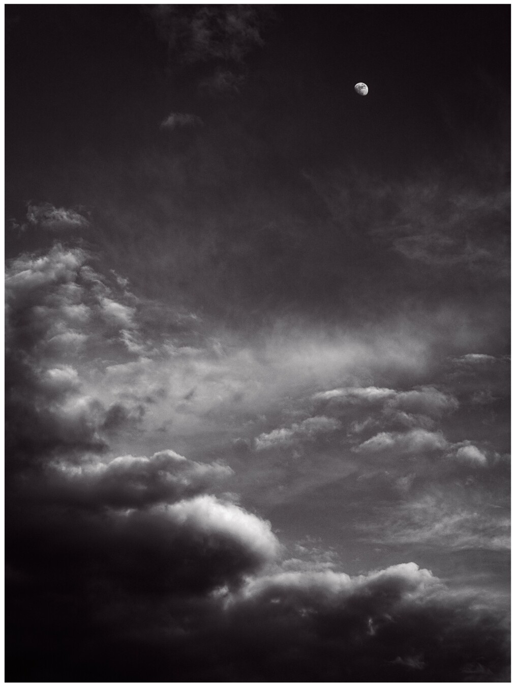 In this black and white photo, a set of clouds frame the left and bottom sides of the photo. The sun highlight their leading edges while the undersides fade into darkness. In the top right corner is the faraway moon.