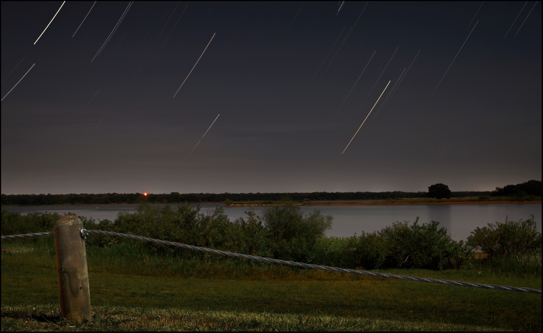 This is a time lapse image illuminated primarily by the light of the moon and stars. The scene is a local lake. In the foreground a barrier cable lit my moonlight stretches from a post on the left to across the right edge of the frame. Behind that, bushes mark the shore of the lake. The lake extends to the far shore and a horizon that bisects the image just below frame center. The night sky fills the rest of the frame.

Numerous stair trails lead up into the sky like an convoy of rocket ships leaving the planet.