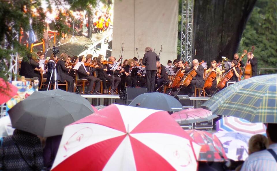 Th bottom part of the picture is a bunch of colorful umbrellas, the closer to the camera is a red and white umbrella. The top part of the picture is the filarmonica on the stage performing.