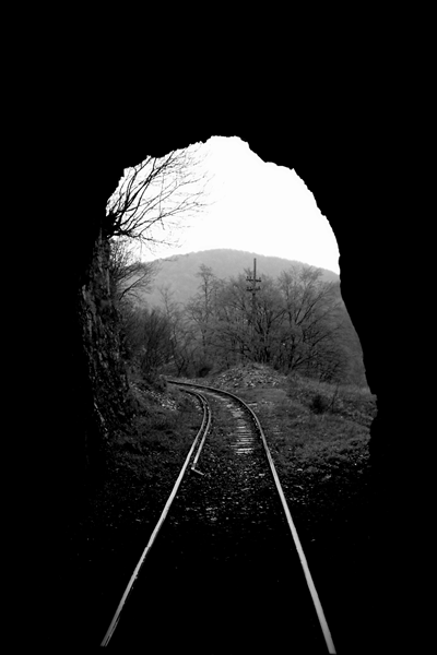 A black and white shot of the tracks being left behind as the train enters a tunnel. The sides of the tunnel look completely black with the opening looking like a portal towards a softly lit landscape of hills and forests. The tracks curve towards the left and vanish,