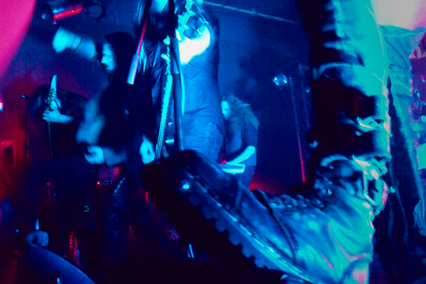The boot of one of the musicians is in the foreground, resting on the speaker, uncomfortably close to the camera. Behind the rest of the band is performing. The photo is very hazy, captured in blue light with some pink accents.