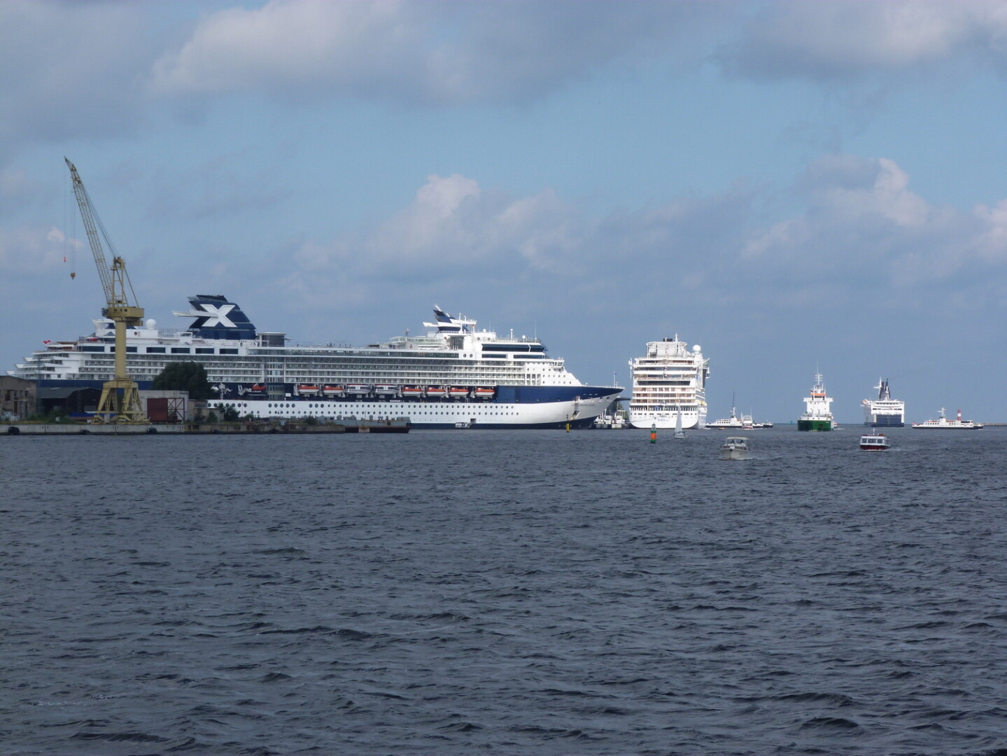 Weißes Kreuzfahrtschiff mit blauem X-Logo am Kai, umgeben von kleineren Booten und einem Kran unter einem bewölkten Himmel.