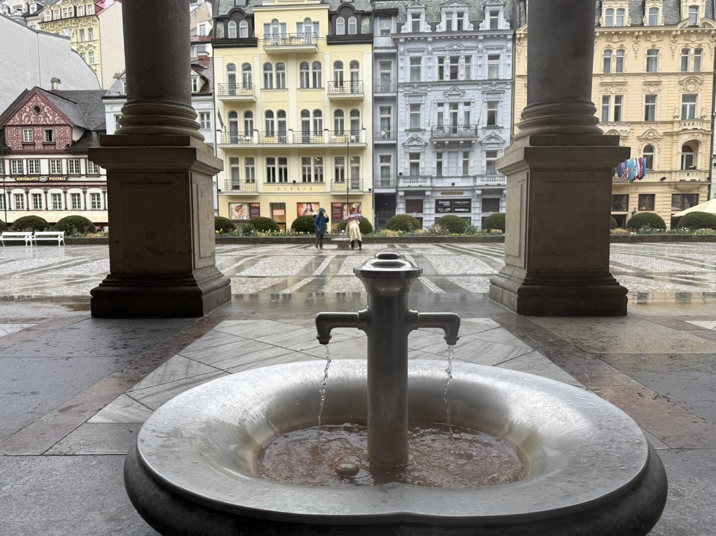 Brunnen mit zwei Wasserhähnen in einem gepflasterten Platz mit bunten historischen Gebäuden im Hintergrund.