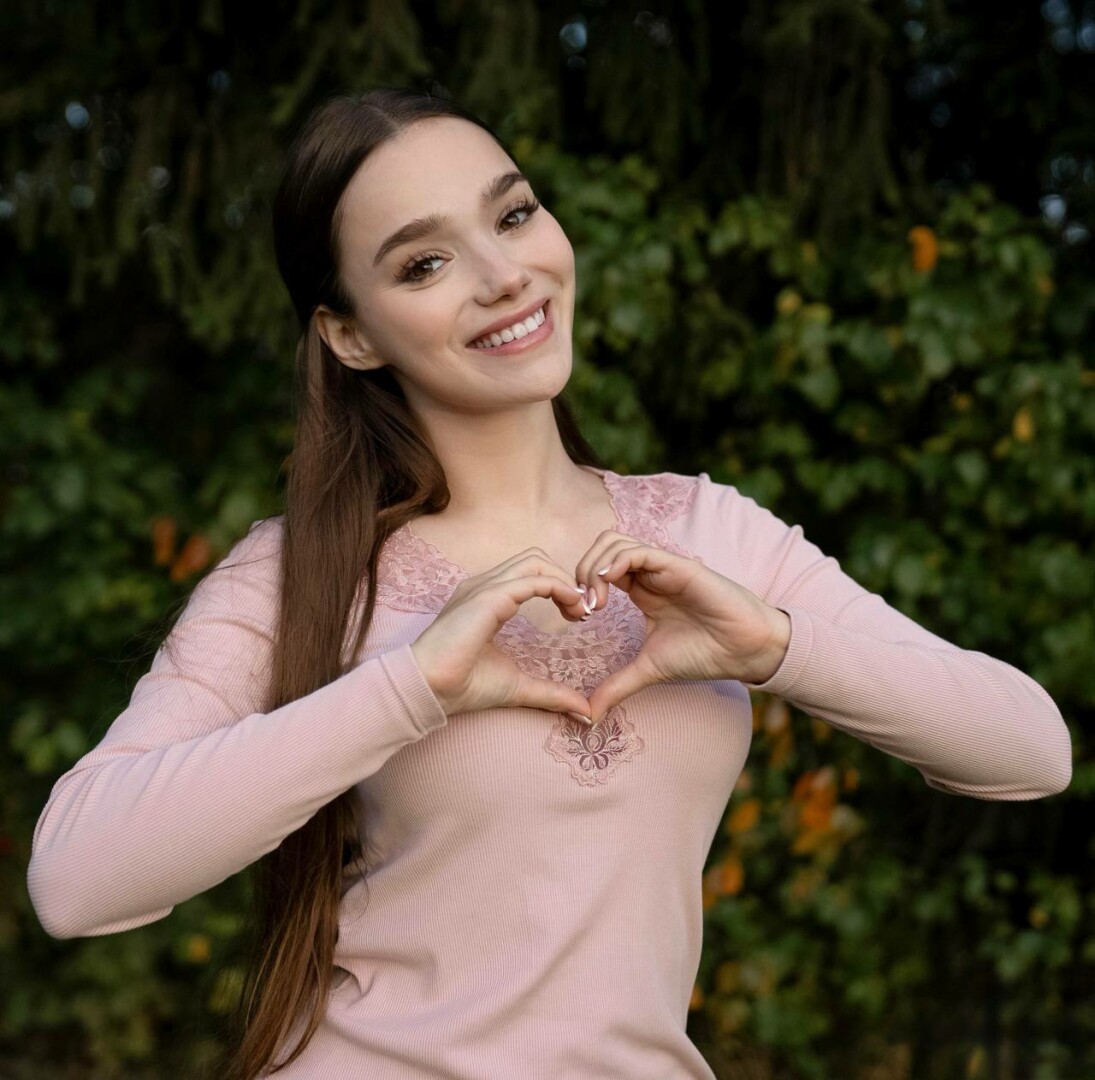 A young woman posing indoors, wearing a matching blue cropped blazer and skirt with fishnet tights. She stands confidently with her hands on her waist in front of a wooden table and an ornate chandelier. Warm lighting enhances the soft-glamour, fashion-focused mood.