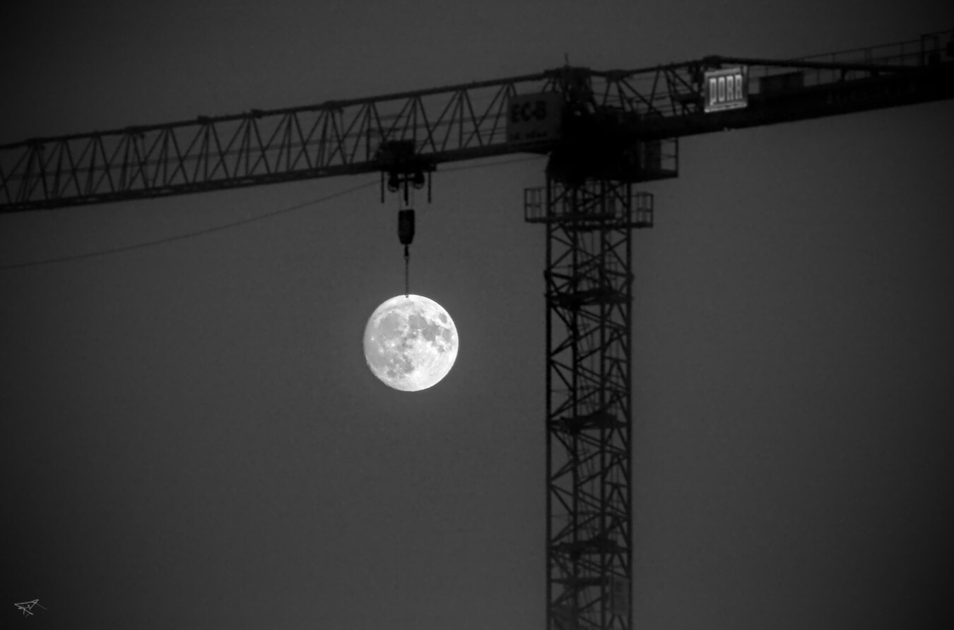 Das Foto zeigt einen Kran in Silhouette vor einem dunklen Himmel. An einem Haken des Krans hängt der volle Mond, der hell und klar leuchtet. Das Bild ist in Schwarz-Weiß gehalten.
 --- 
The photograph shows the silhouette of a crane against a dark sky. The full moon, shining brightly and clearly, hangs from one of the crane’s hooks. The image is in black and white.