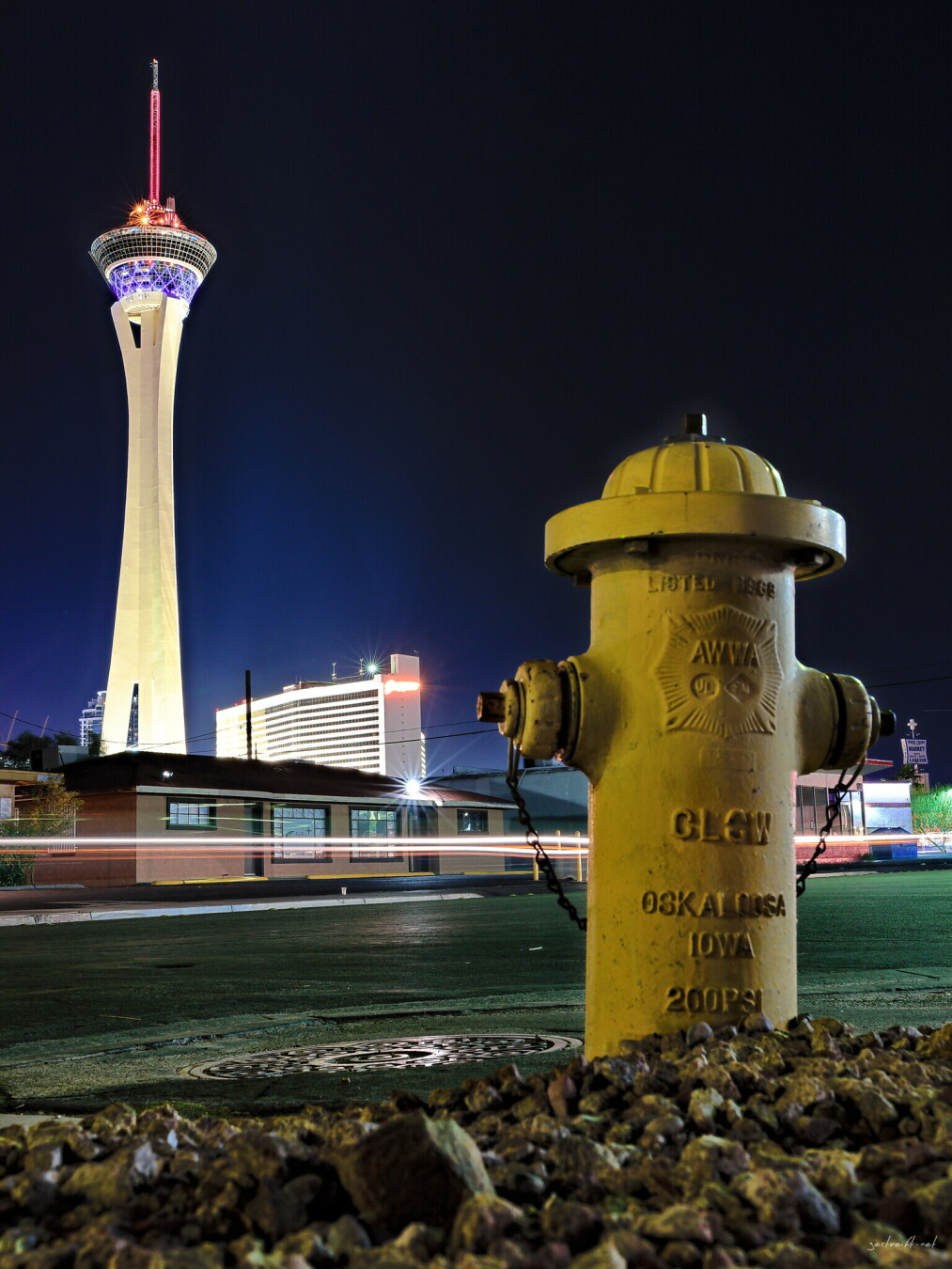 A low-angle night photograph in Las Vegas with a bright yellow fire hydrant in the foreground on a patch of gravel. Behind it, an asphalt street shows light trails from passing cars. In the distance, the Strat tower is illuminated with red and purple lights against a dark sky. Other city lights are visible on nearby buildings. The hydrant text is readable: "AWWA," "GLOW," "OSKALOOSA," "IOWA," and "200PSI."
