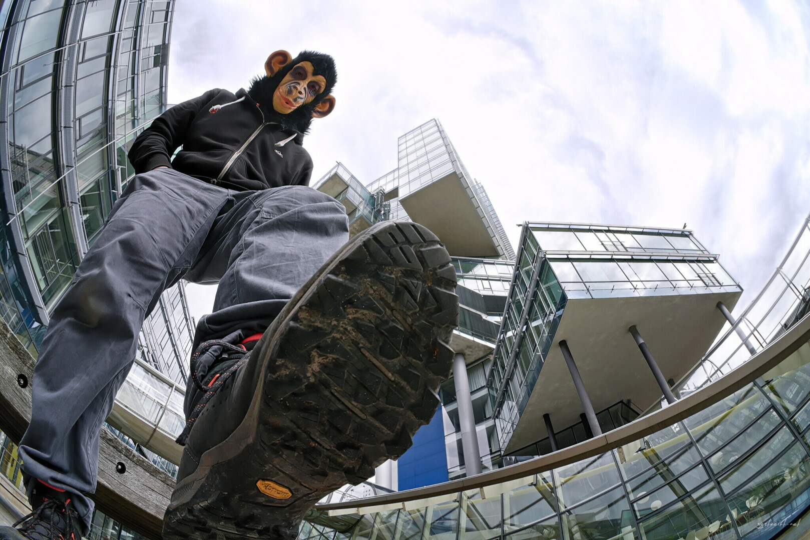 A low-angle, fisheye shot of a person wearing a chimpanzee mask standing dramatically over the camera. They wear a black hoodie, grey pants, and dark boots, stepping one boot close to the lens. Modern, geometric glass architecture and an overcast sky tower in the background.