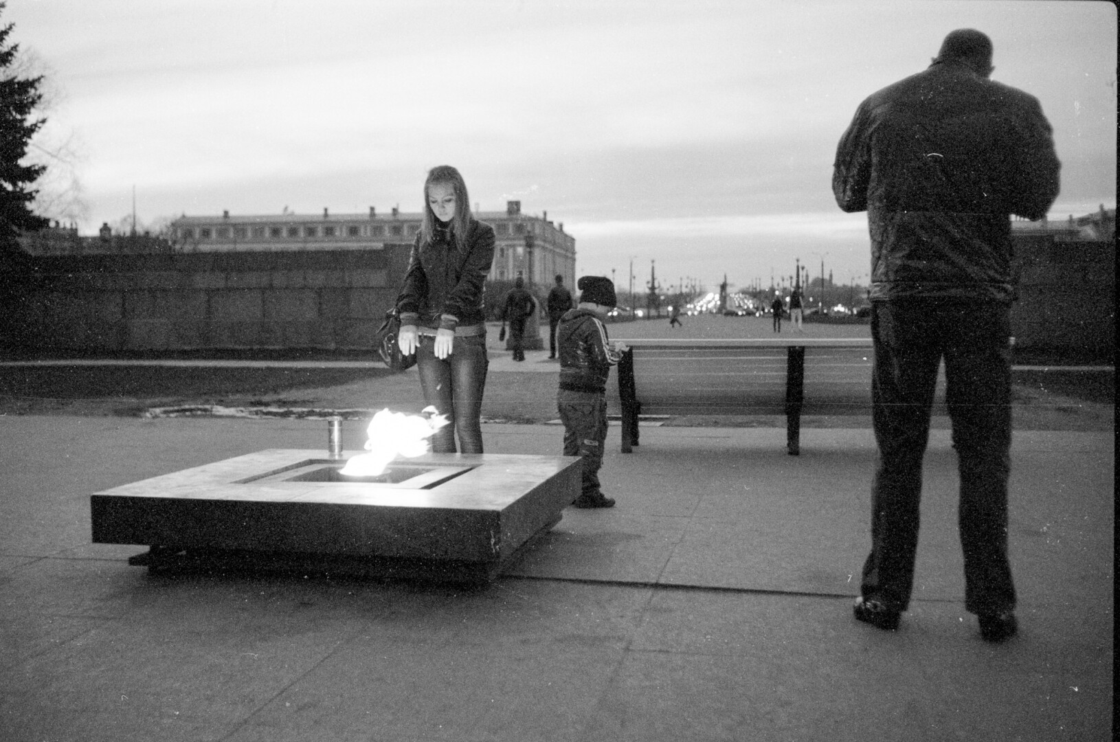 A woman, a child, and a tall man is standing next to a memorial with a flame. The woman is warming herself on the flame.