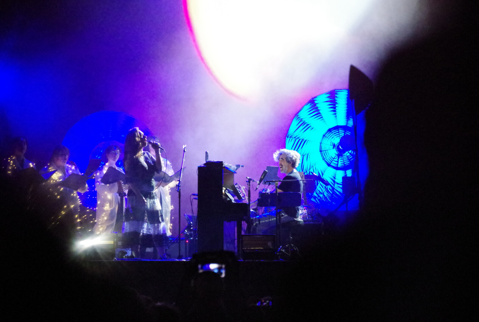 A pianist is performing on stage and singing while seated. He is backlit with his side facing the audience. A pair of singers are in front of him.