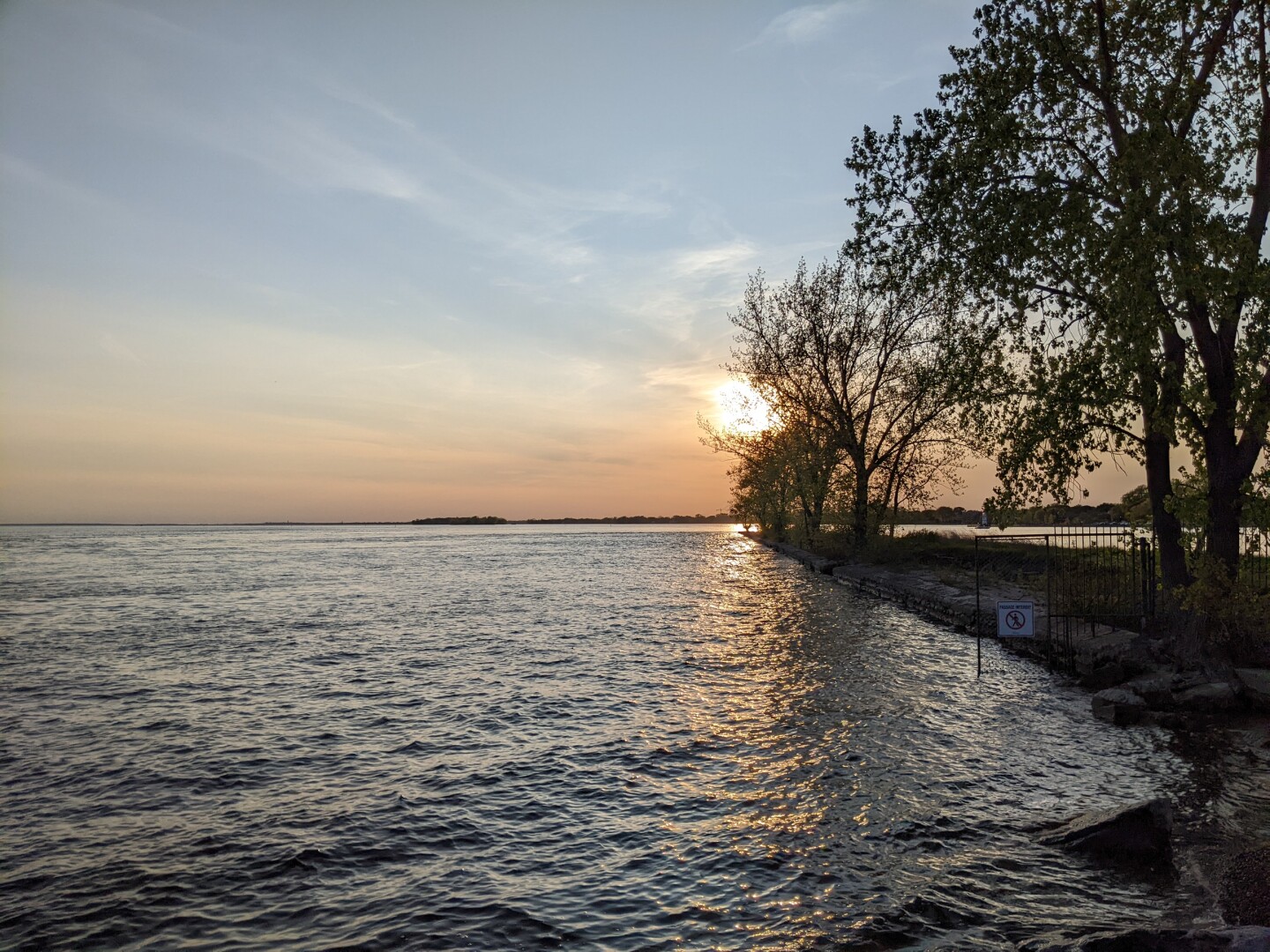 The sun setting on a body of water. A strip of land with a few trees juts out on the right. In the far distance, land can be seen.