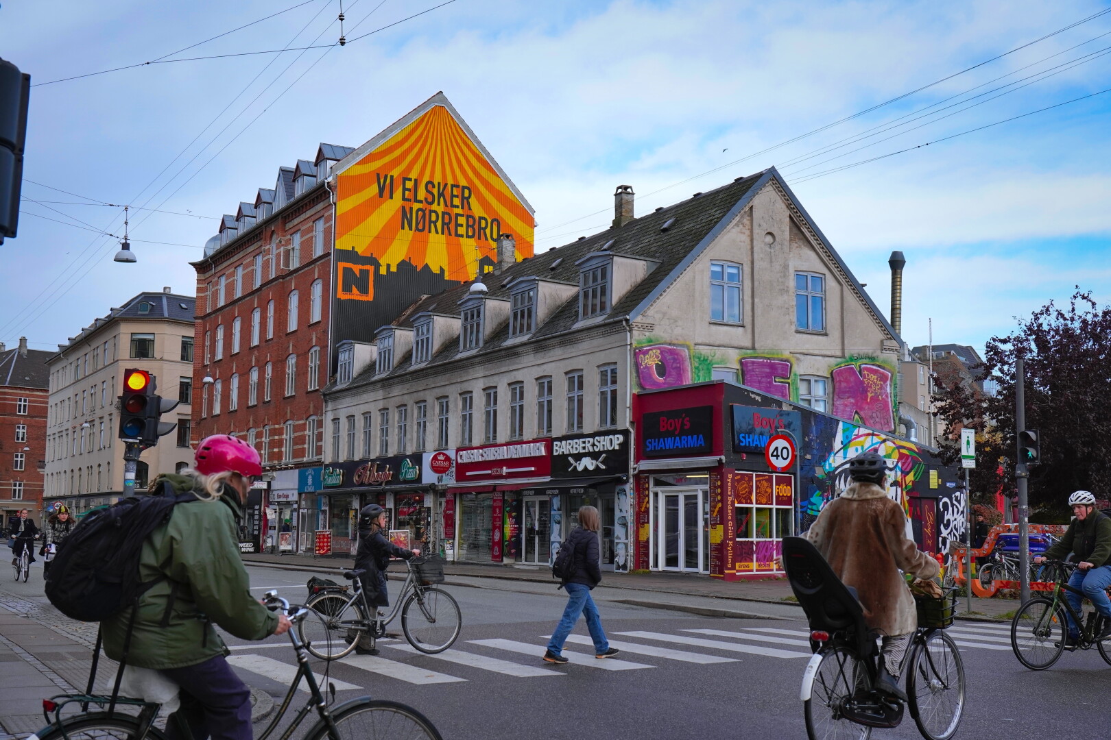 A picture of a cross work in Nørrebro, Copenhagen. Many people cross by bike and by foot. In the background is a house with a mural. The mural contains the colours black, yellow and orange. The skyline of norrebro is painted in black, the sky is made of yellow and orange stripes.