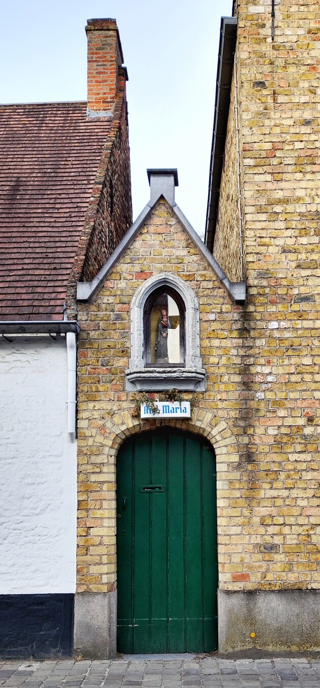 A small, narrow green wooden door nestled between two taller brick buildings, with a statue of the Virgin Mary and the inscription ‘Ave Maria’ in blue Gothic letters.